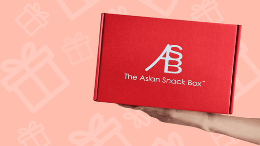 Hand holding a red box labeled 'The Asian Snack Box' with a gift-themed background.